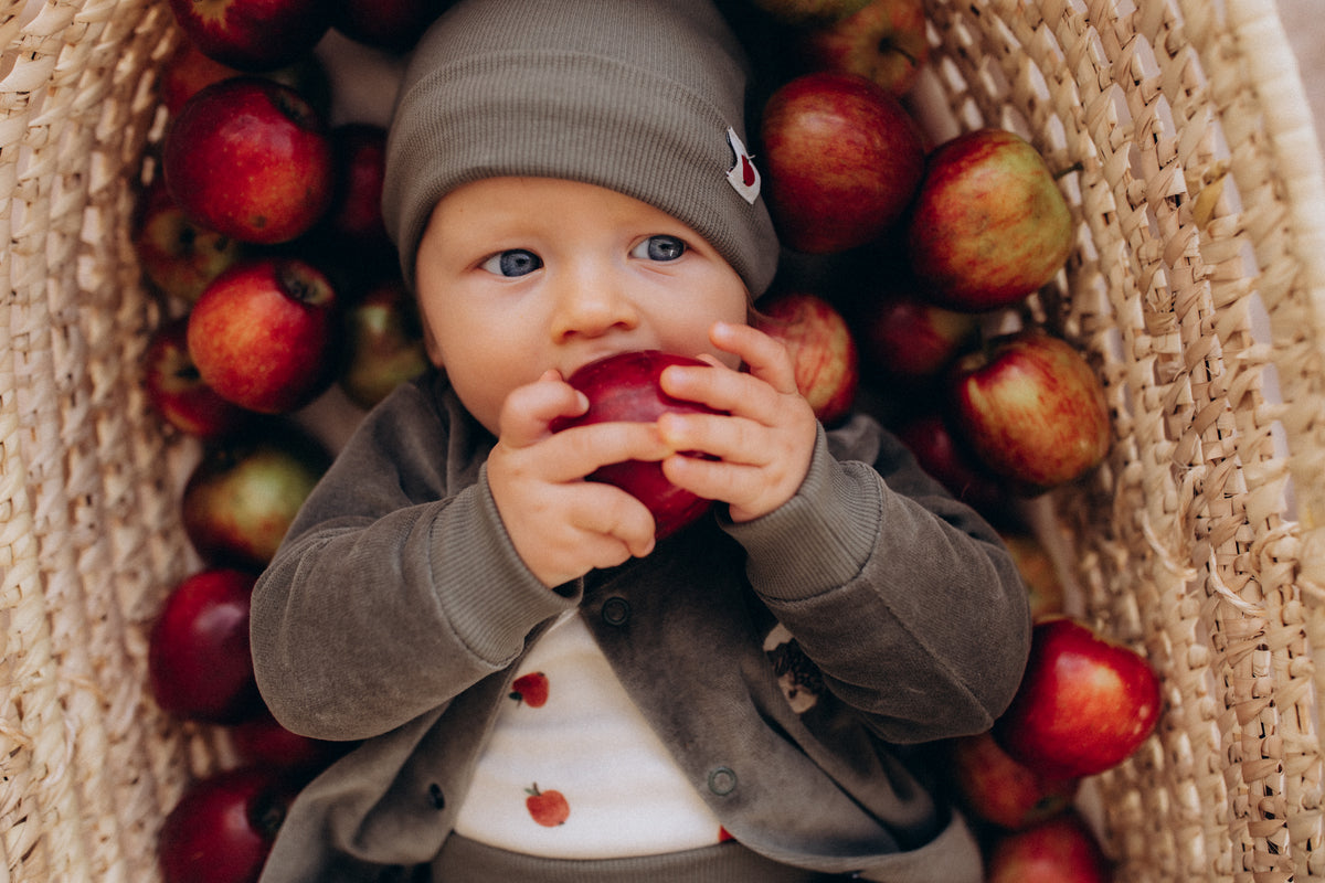 Baby in a knitted beanie sitting outdoors in a basket of apples