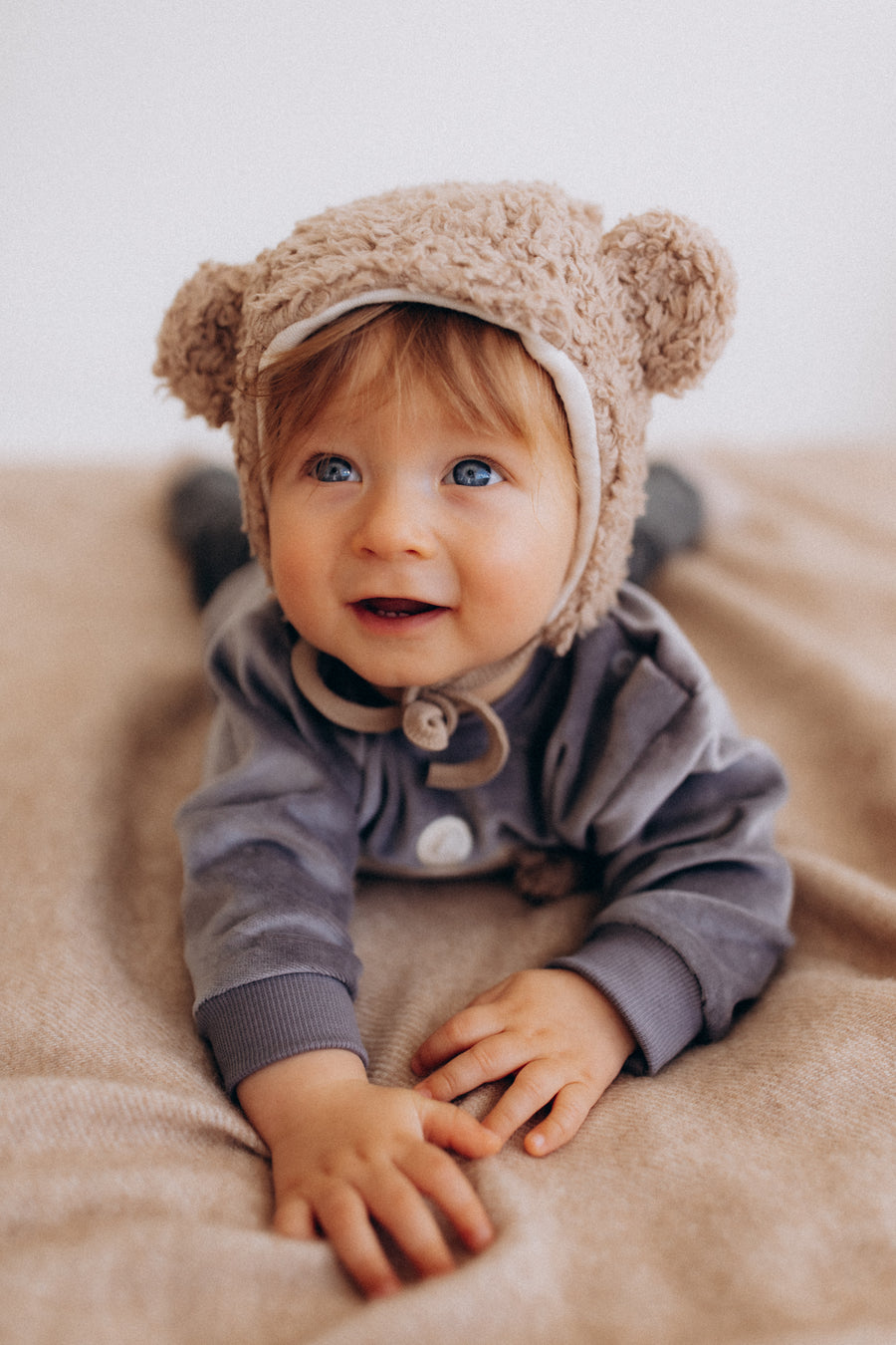 Baby in a soft bear-ear bonnet resting on a tan blanket